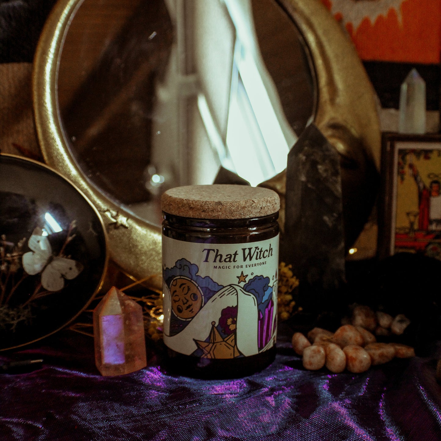 Candle labeled 'That Witch' on a purple fabric with decorative items in the background