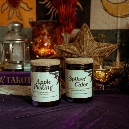 Two jars labeled 'Apple Picking' and 'Spiced Cider' on a purple surface with decorative items in the background.